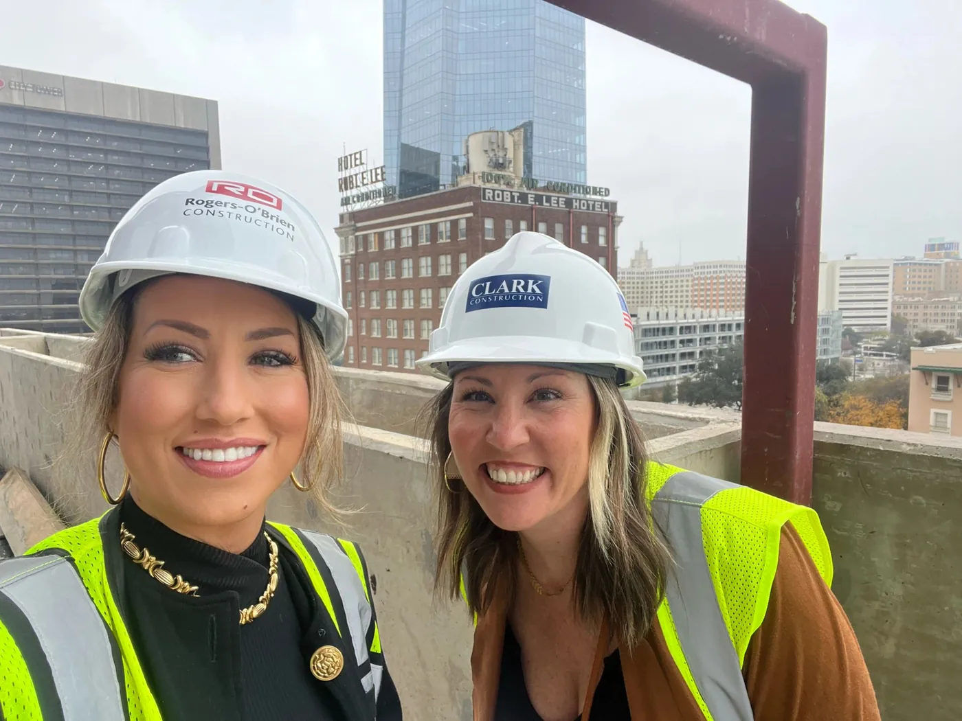Sarah Loredo and Amanda Mercer in hard hats and safety vests on a rooftop job site in downtown San Antonio