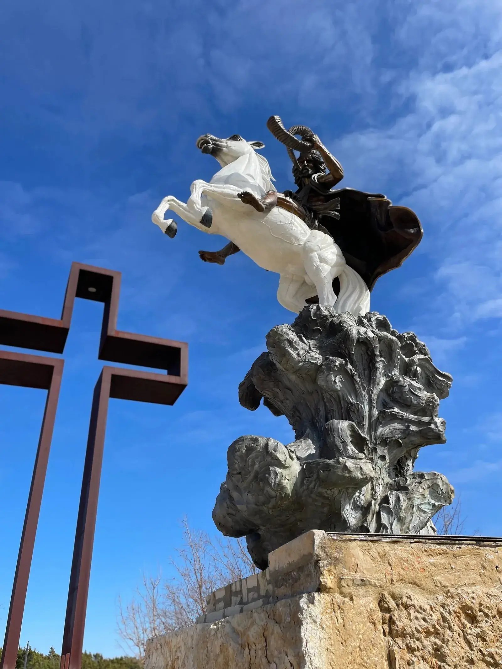 Bronze-and-white horse and rider sculpture on a limestone base beside a tall steel cross under a blue Hill Country sky